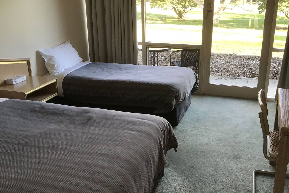 Ein Hotelzimmer mit zwei Betten, einem Fenster mit Blick auf einen Park und einem kleinen Tisch mit einem Stuhl.
