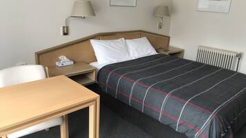 A hotel room with a bed, a desk, two bedside lamps, a radiator, and two framed artworks on the wall.