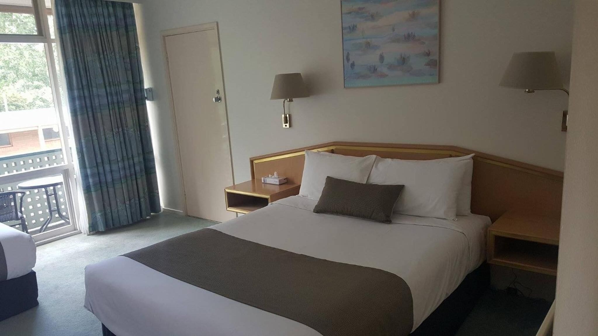 A hotel room with a large bed, two bedside tables, a nightstand, a window with curtains, and a painting on the wall.