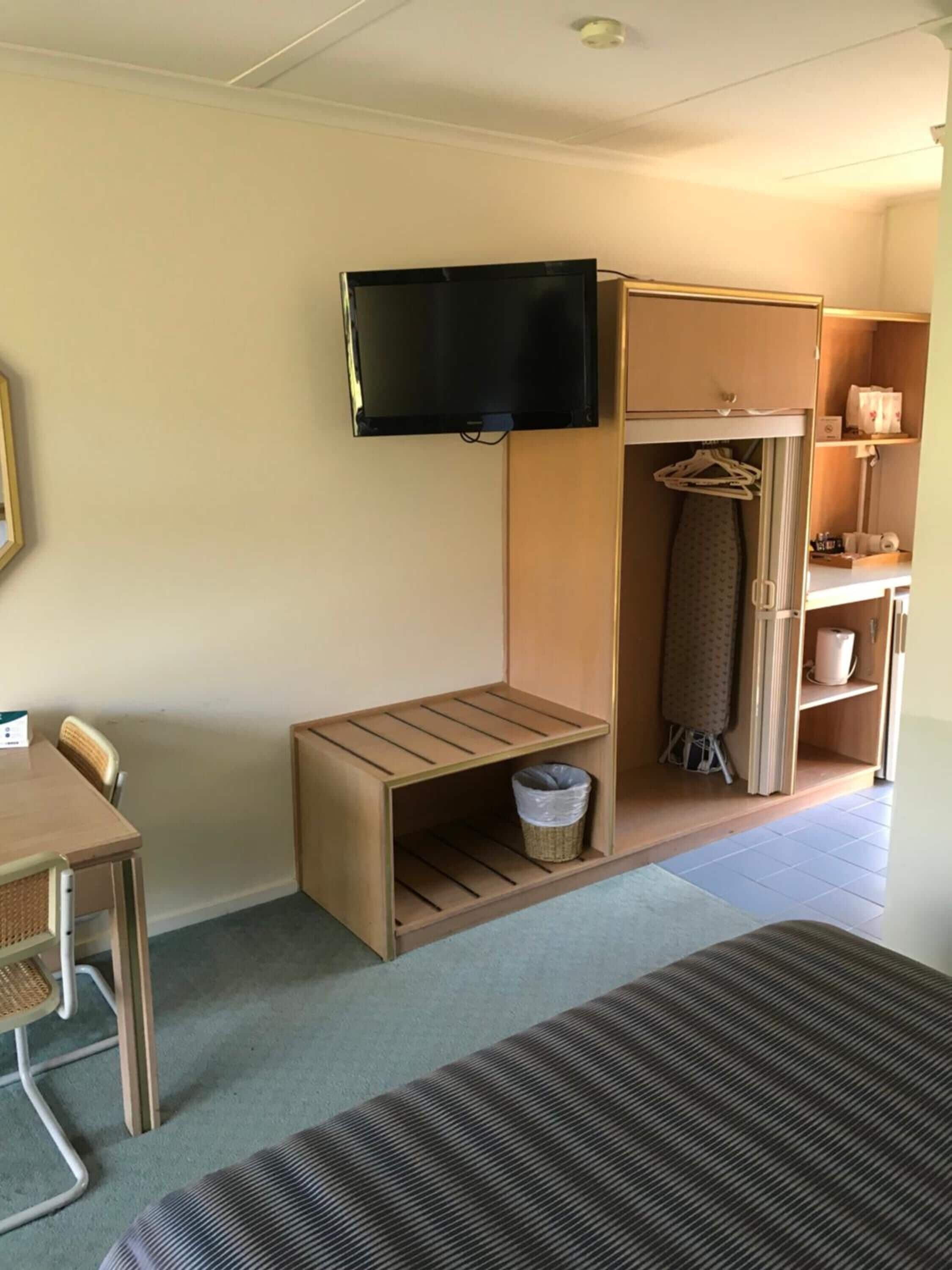 A compact hotel room with a bed, a small desk, a television mounted on the wall, a wardrobe, and a closet with hanging clothes.