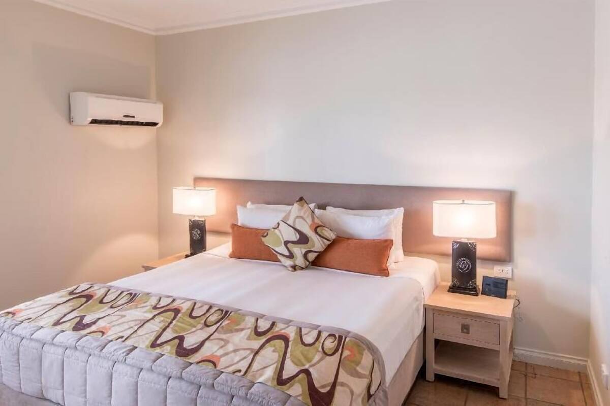 Una habitación de hotel con una cama, dos lámparas de noche, un mesita de noche y un equipo de aire acondicionado en la pared.