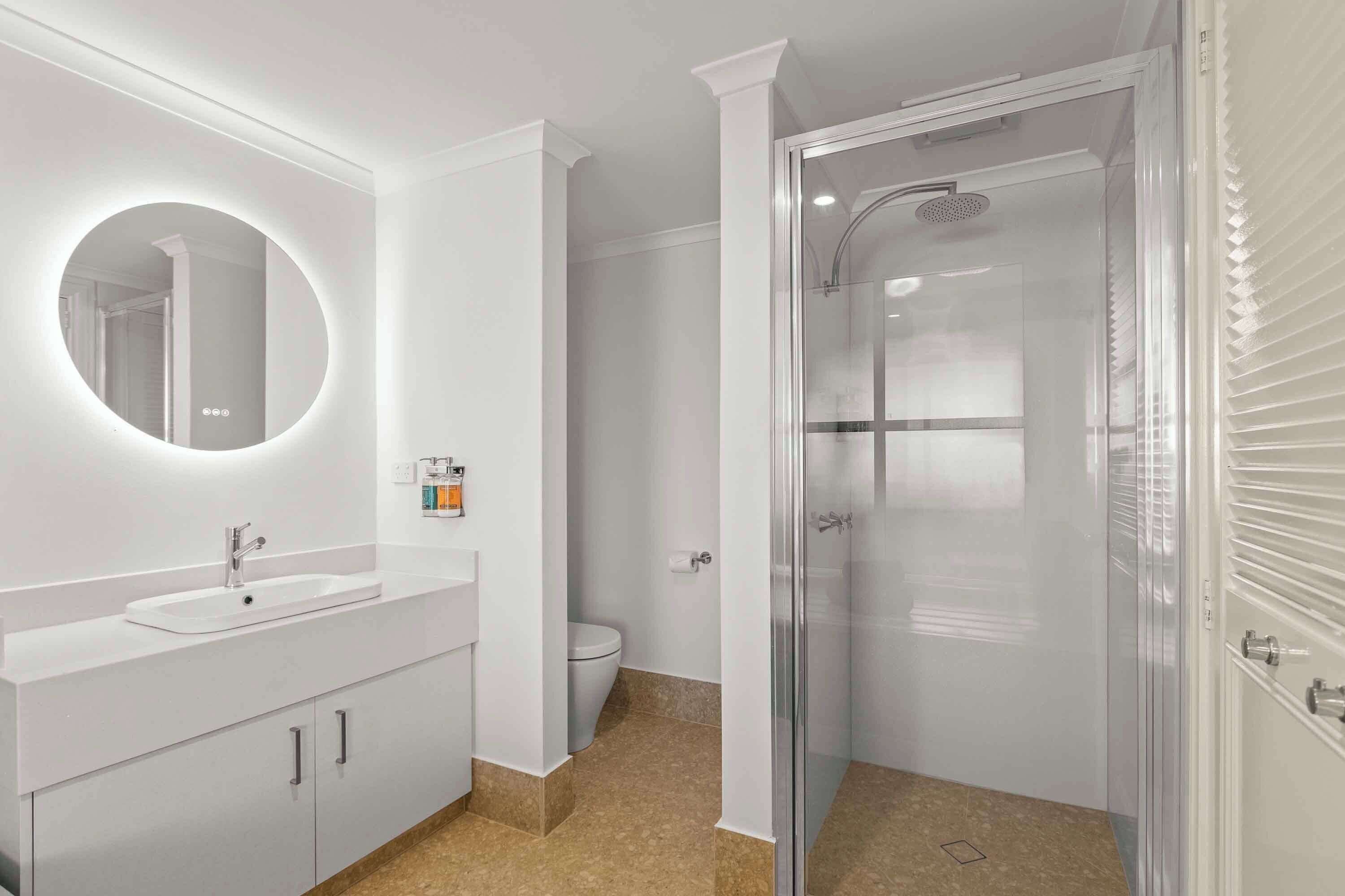 Un baño moderno con un espejo redondo, un lavamanos y una ducha con mampara de vidrio.