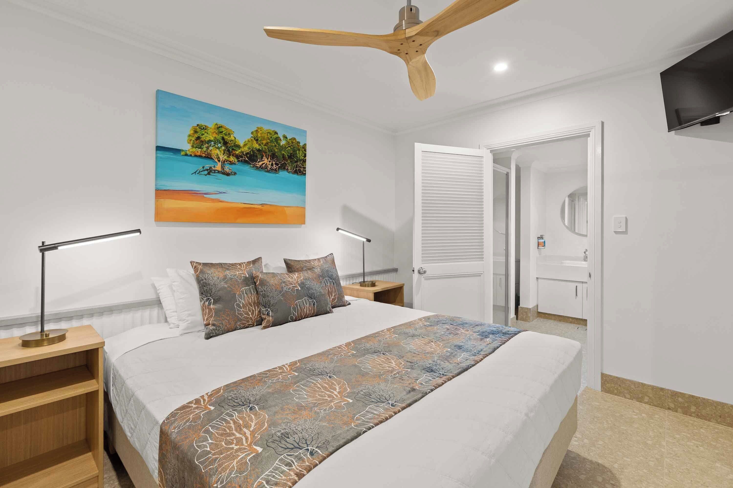 Une chambre à coucher avec un lit, un tableau représentant une plage, un ventilateur de plafond, une télévision et une salle de bain avec un lavabo et un miroir.