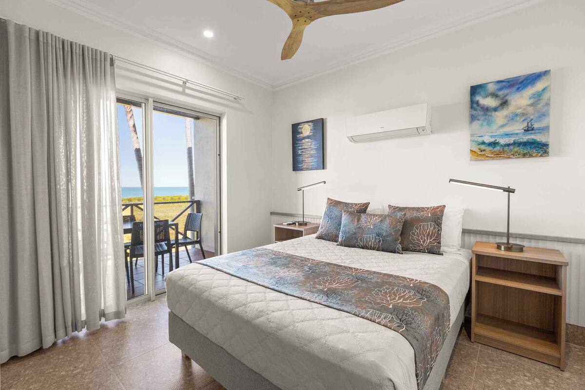 Un dormitorio con una cama grande, una mesita de noche de madera, un ventilador de techo y vista al mar a través de una puerta corrediza.
