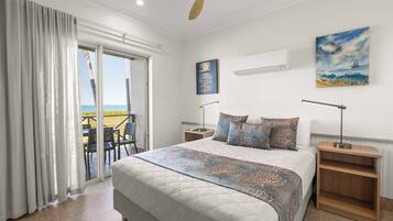 Quarto com uma cama grande, mesa de cabeceira de madeira, ventilador de teto e vista para o mar através de uma porta deslizante.