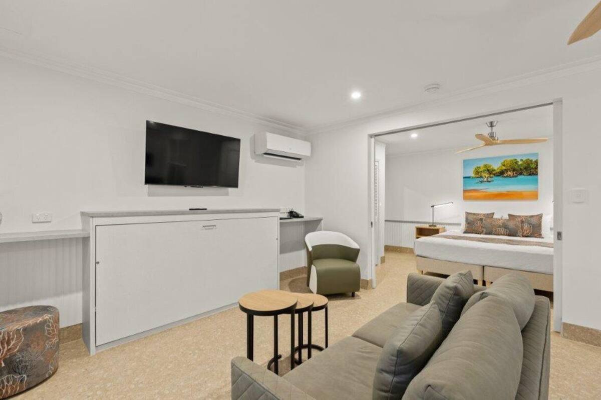 Una sala moderna con un sofá gris, una mesita redonda, un televisor montado en la pared y un cuadro con una playa.