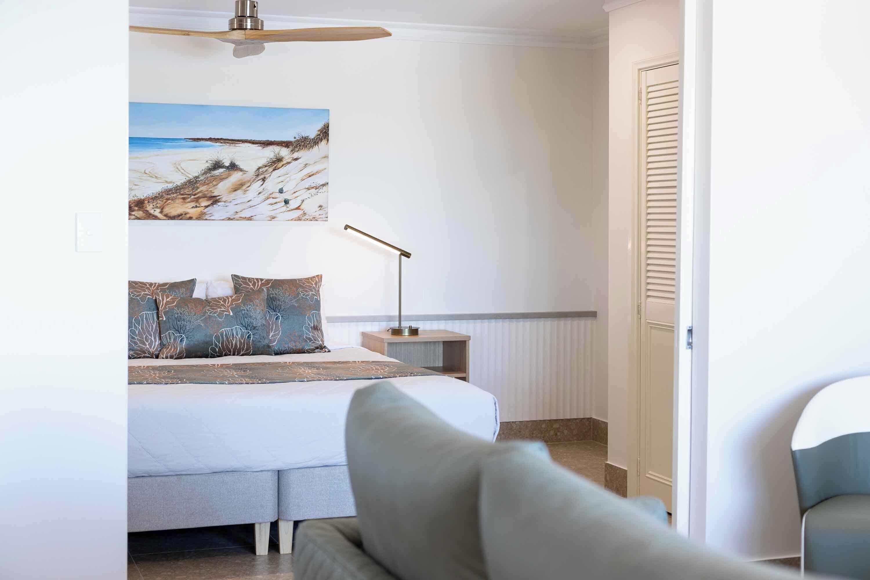 A bedroom with a bed, a nightstand, a lamp, and a painting of a beach scene.