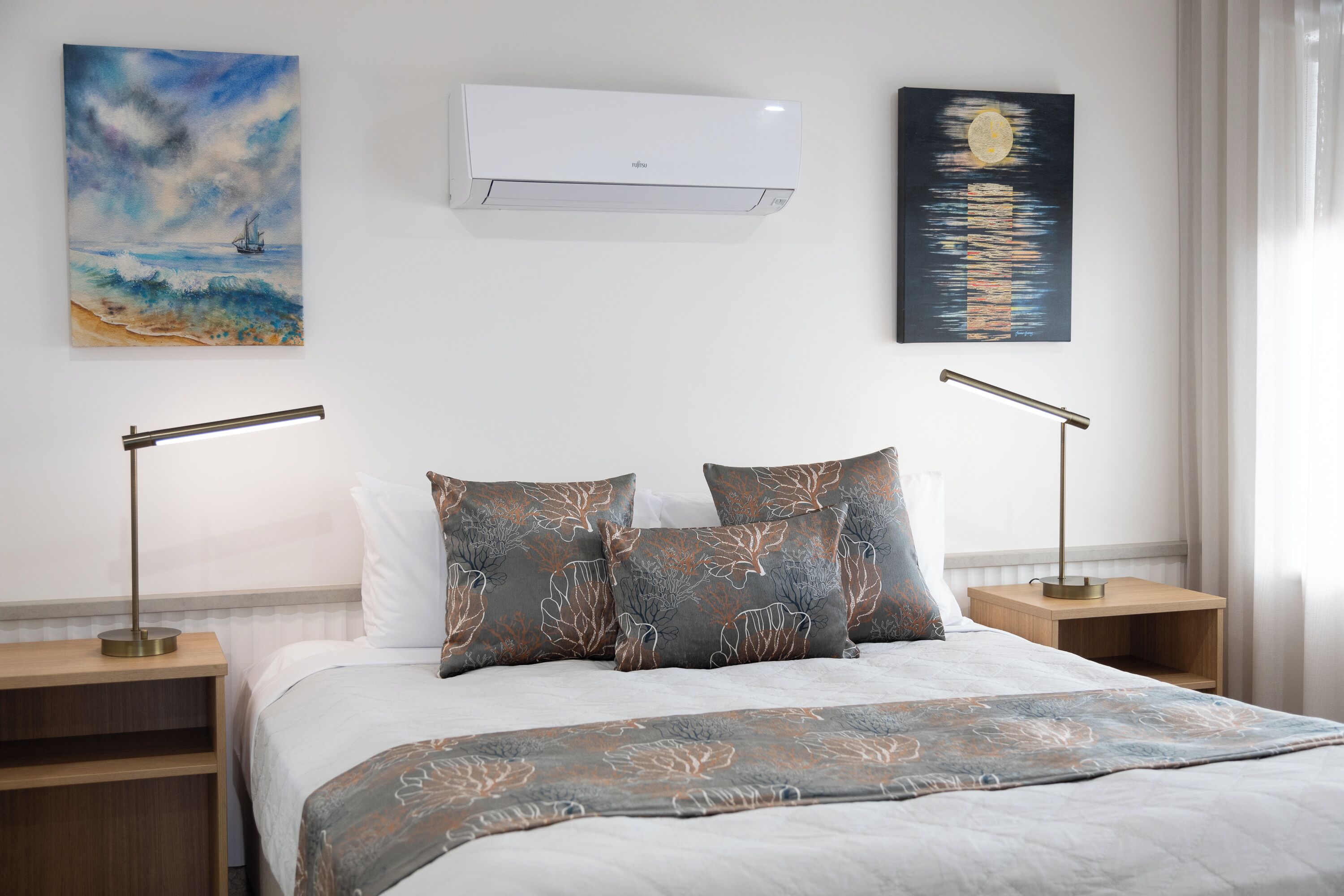 Una cama bien hecha con un edredón estampado con motivos de hojas, dos mesitas de noche con lámparas, un aire acondicionado instalado en la pared y dos cuadros.