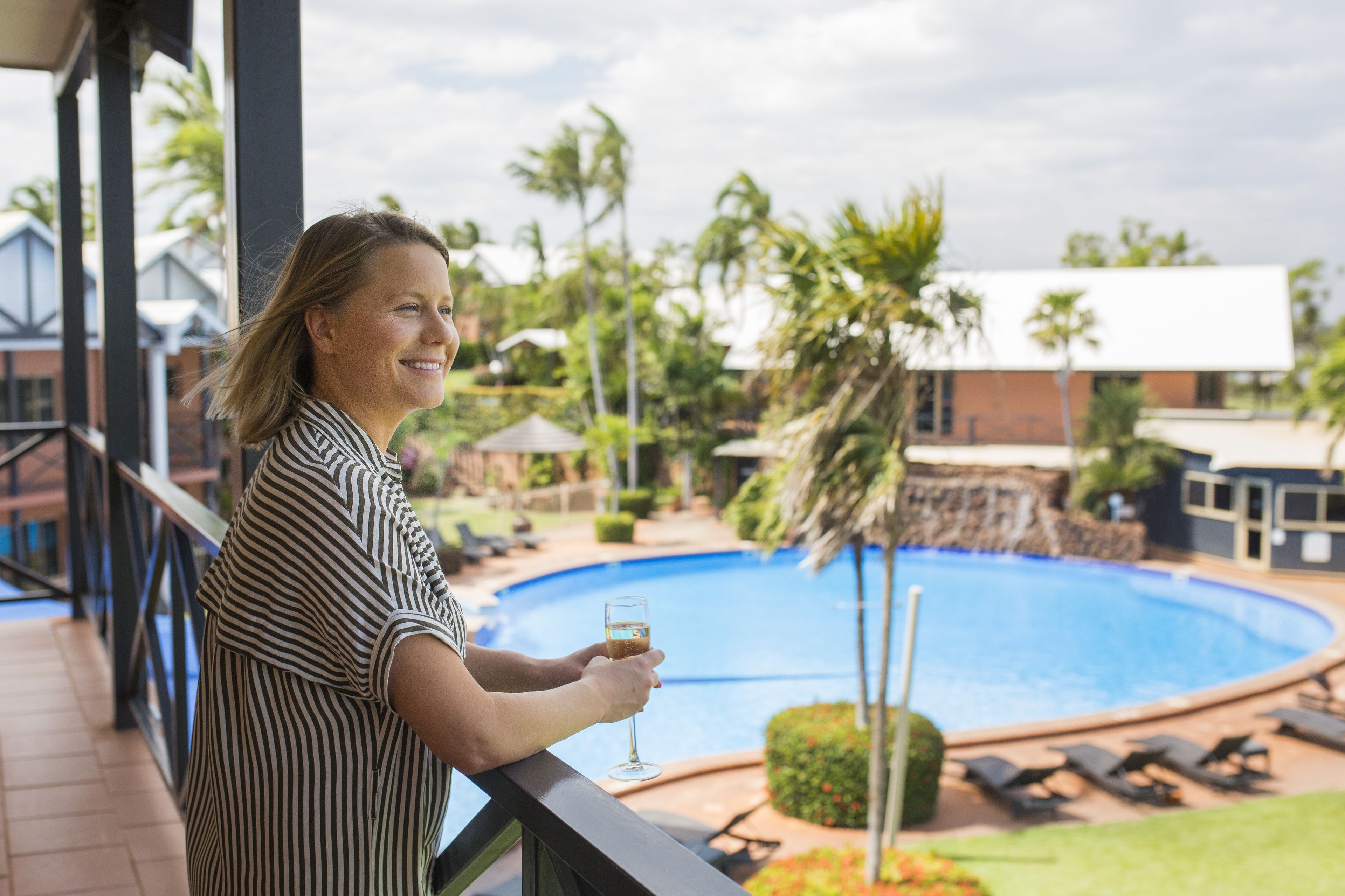 A person holding a glass of drink on a balcony overlooking a swimming pool with lounge chairs and palm trees.