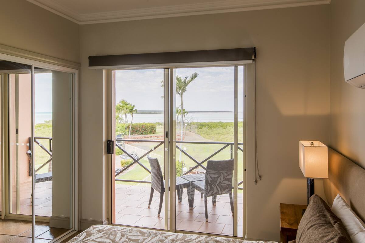 Habitación de hotel con cama, mesita de noche, lámpara y vistas a un patio con sillas y una mesa.