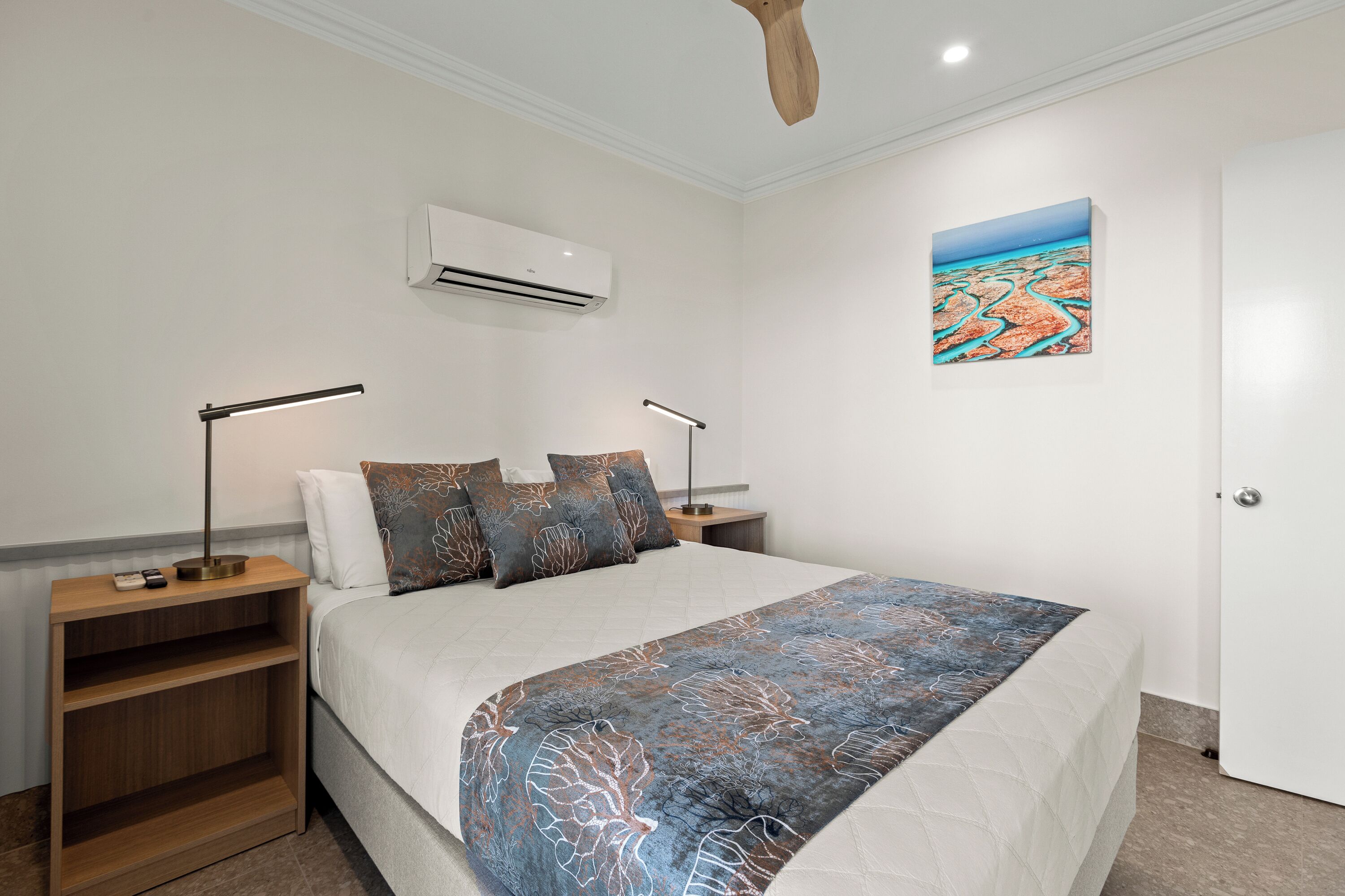 A modern bedroom with a large bed, two bedside tables, a ceiling fan, an air conditioning unit, and a painting on the wall.