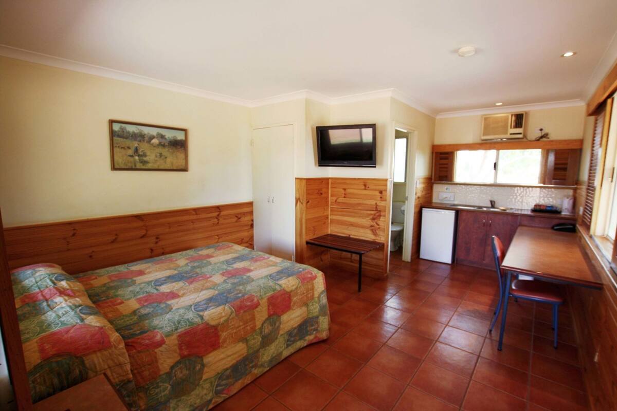 Una habitación compacta con cama, una mesita, televisión y kitchenette.