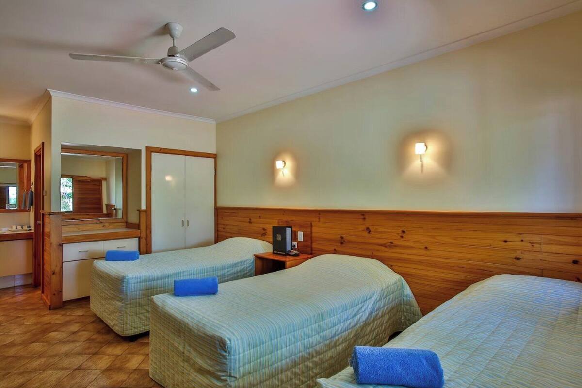 Habitación de hotel con dos camas individuales, cabecera de madera, ventilador de techo, espejo, armario y una mesita con lámpara.