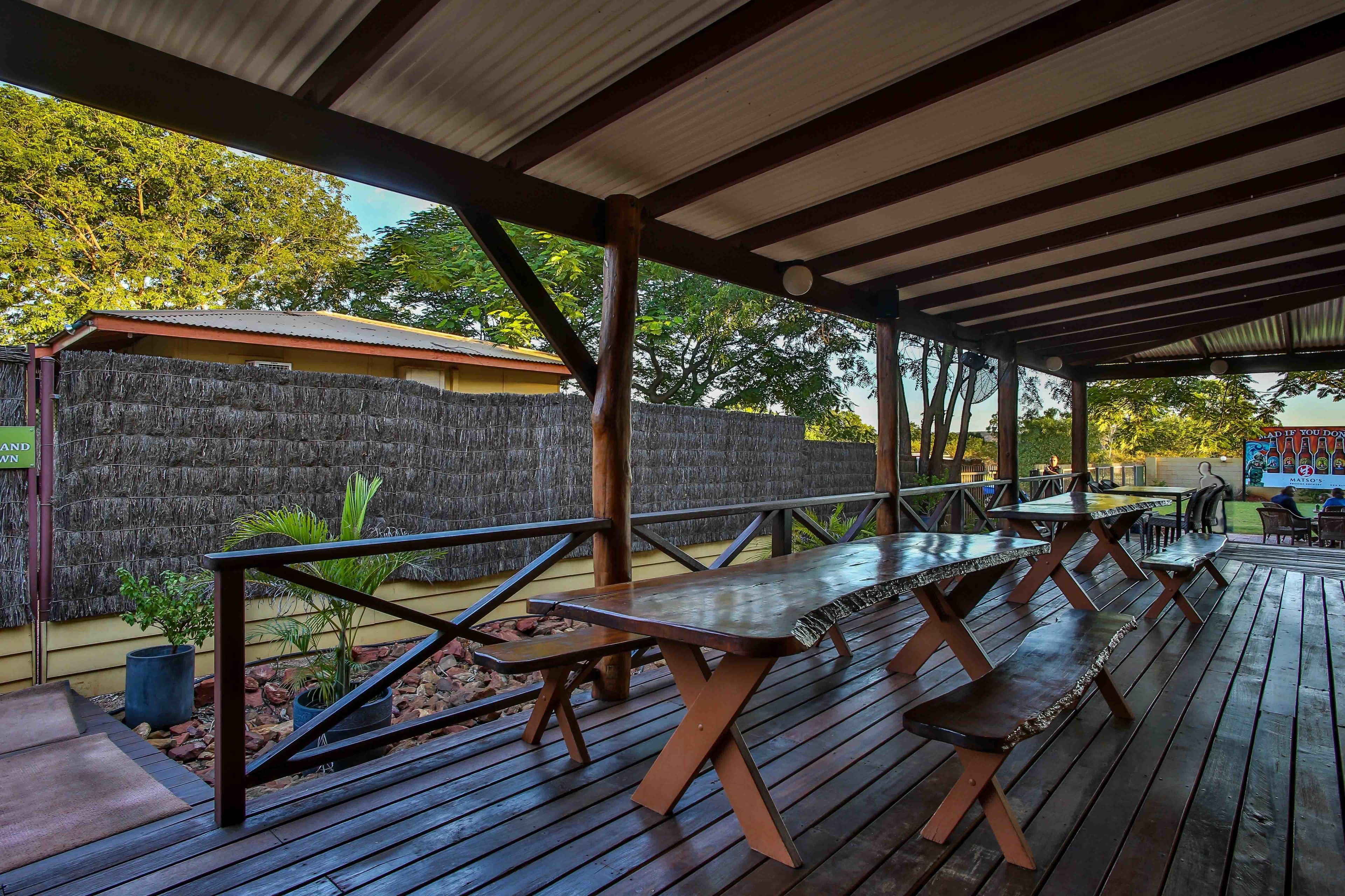An outdoor patio with wooden tables and benches, a covered area with a metal roof, and a stone wall in the background.