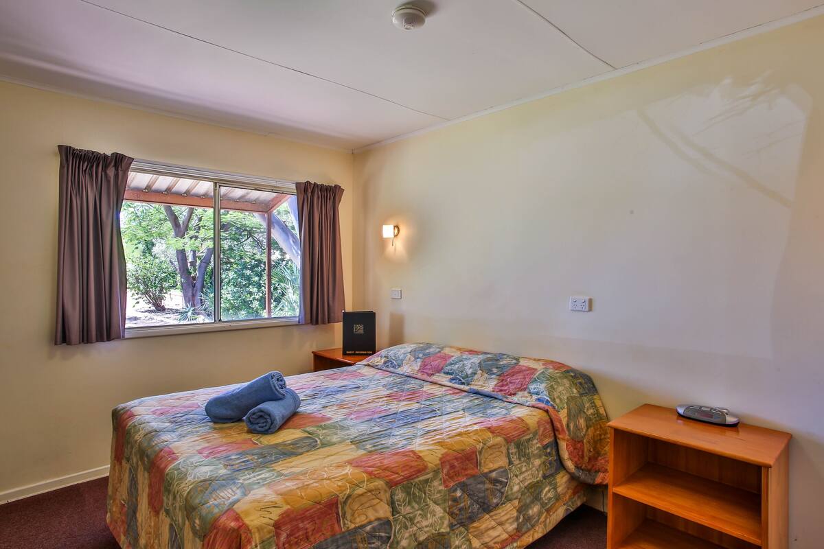 Un dormitorio con cama, ventana con cortinas, mesita de noche con control remoto y una lámpara fijada a la pared.