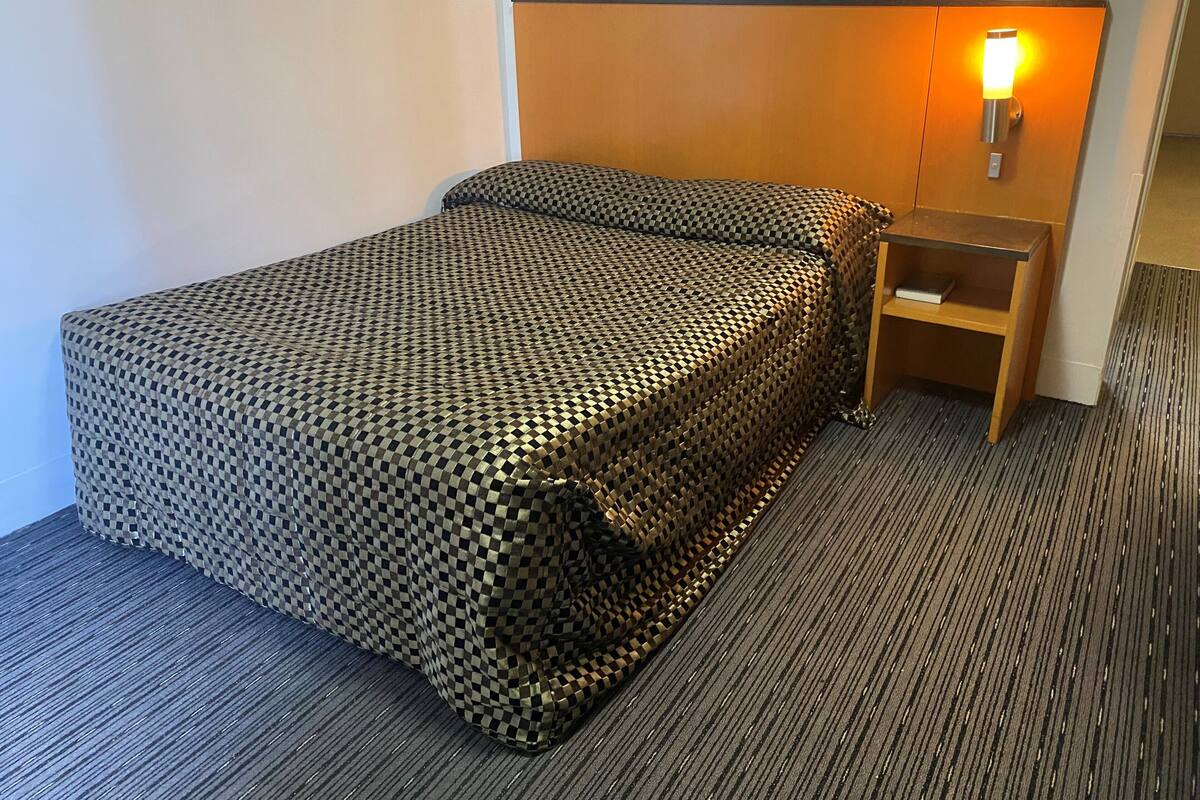 Habitación de hotel con colcha a cuadros, cabecera de madera, mesita de noche con lámpara y suelo alfombrado.