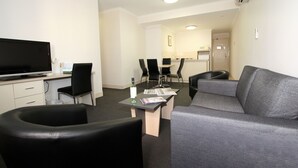 A modern hotel room with a flat-screen TV, a grey sofa, black chairs, a small table with magazines, and a kitchenette in the background. - Comfort Inn & Suites Goodearth Perth (Perth)