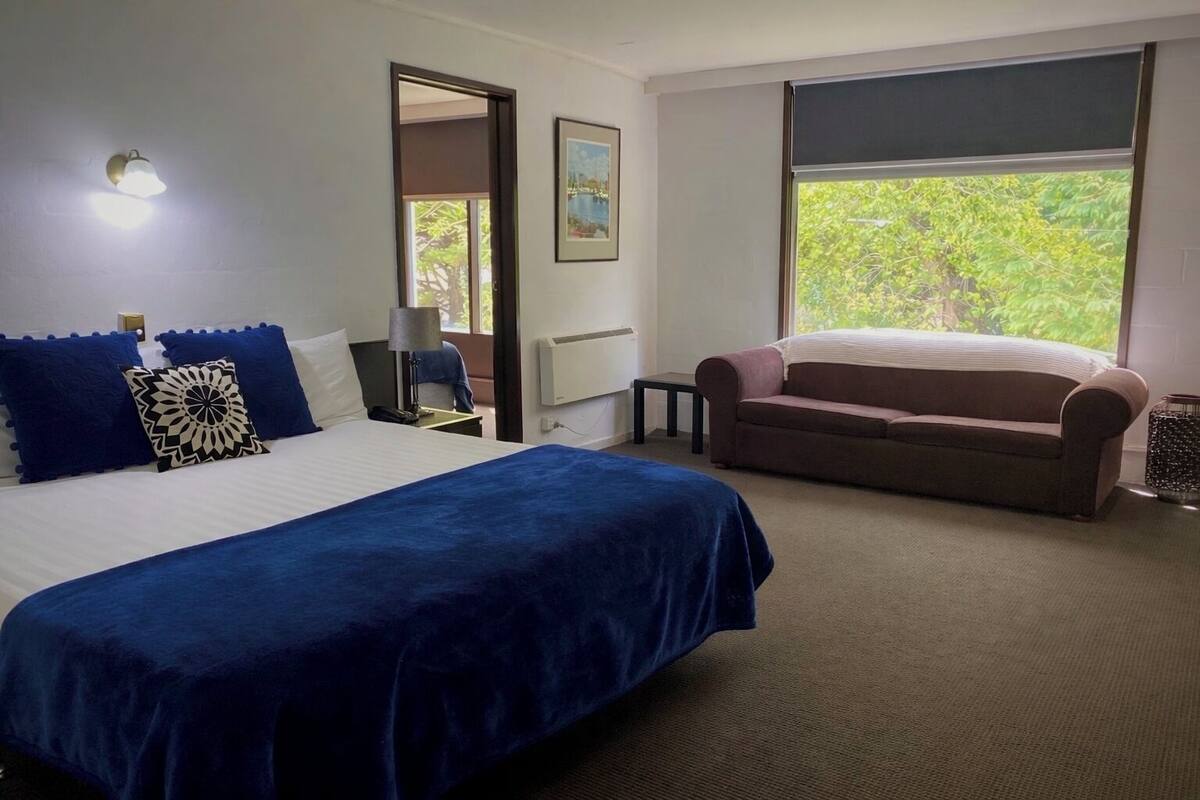 A hotel room with a bed, a sofa, a nightstand, a lamp, a picture on the wall, and a window with a view of greenery.