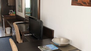 A compact hotel room with a dark wood dining table, beige upholstered chairs, a flat-screen TV, a bowl, and a small trash bin. - West Coaster Motel (Queenstown)