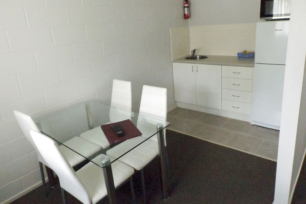 A small, modern kitchen with white cabinets, a microwave, and a sink. There is a glass table with two white chairs and a remote control on it.