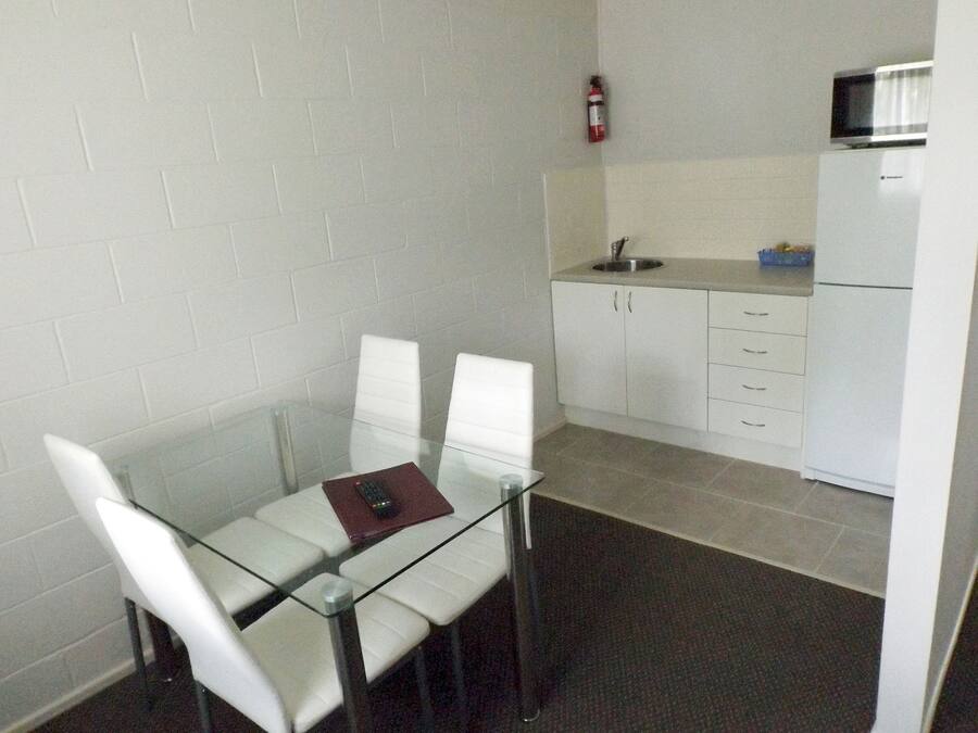A small, modern kitchen with white cabinets, a microwave, and a sink. There is a glass table with two white chairs and a remote control on it.