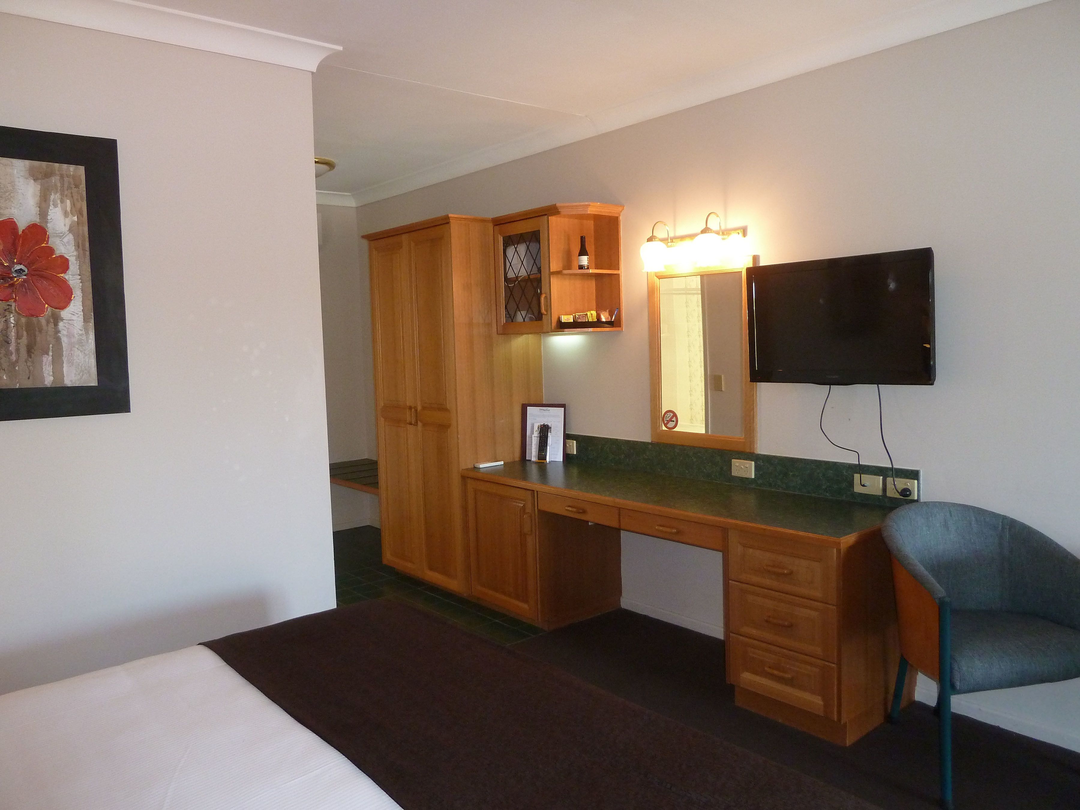 Habitación de hotel con escritorio de madera, una silla, televisor, espejo con luces y un cuadro en la pared.