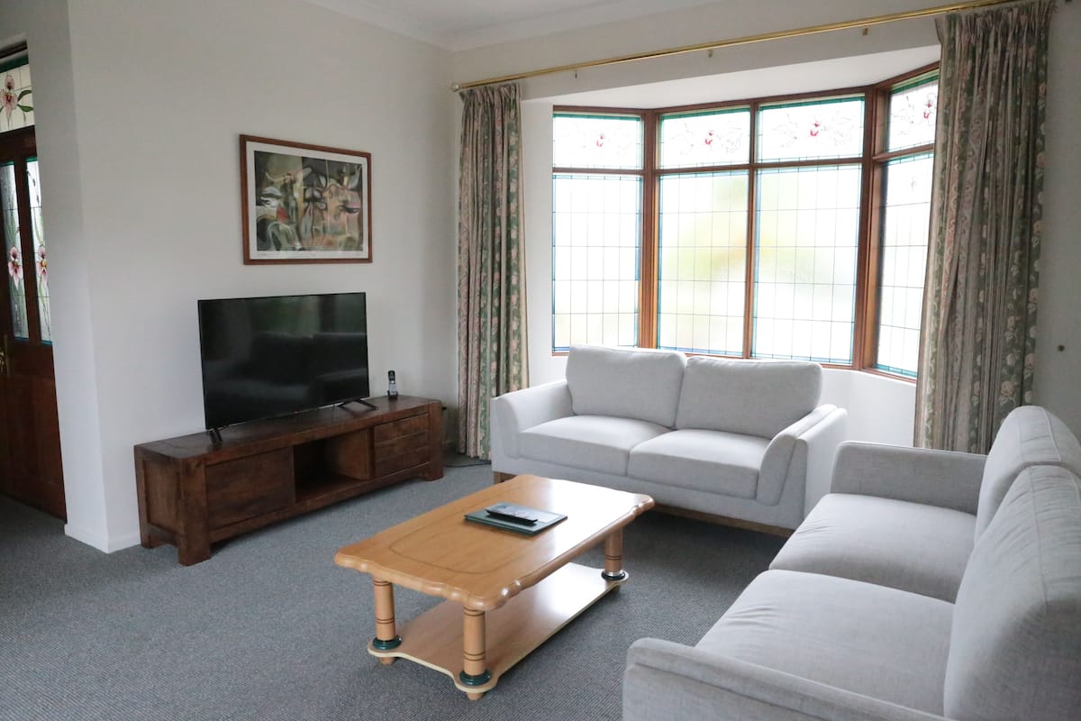 A living room with a flat-screen TV on a wooden stand, a light-colored sofa, a wooden coffee table, and a large window with curtains.