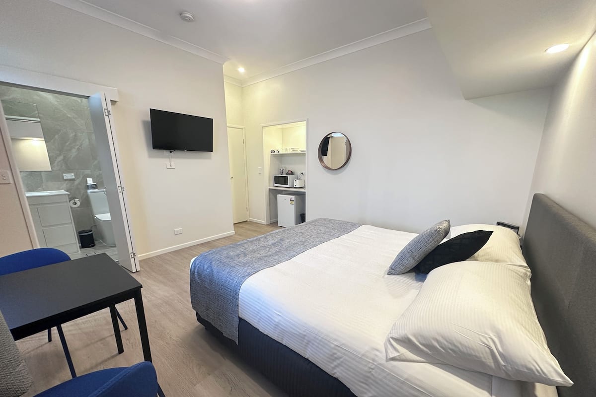 A hotel room with a bed, a desk, a chair, a TV, a small kitchenette, and a bathroom.