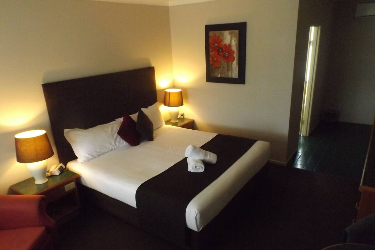 A neatly made bed with white linens, two bedside lamps, a red chair, and a painting on the wall.
