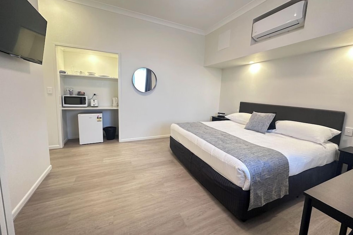 A bedroom with a bed, bedside table, and a small kitchenette area with a microwave and refrigerator.