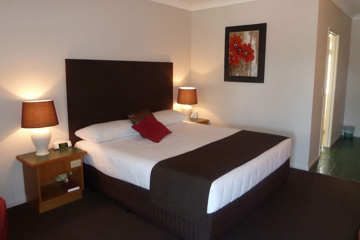 A neatly made bed with a brown headboard, two bedside lamps, a small wooden nightstand with a book and a phone, and a framed artwork on the wall.