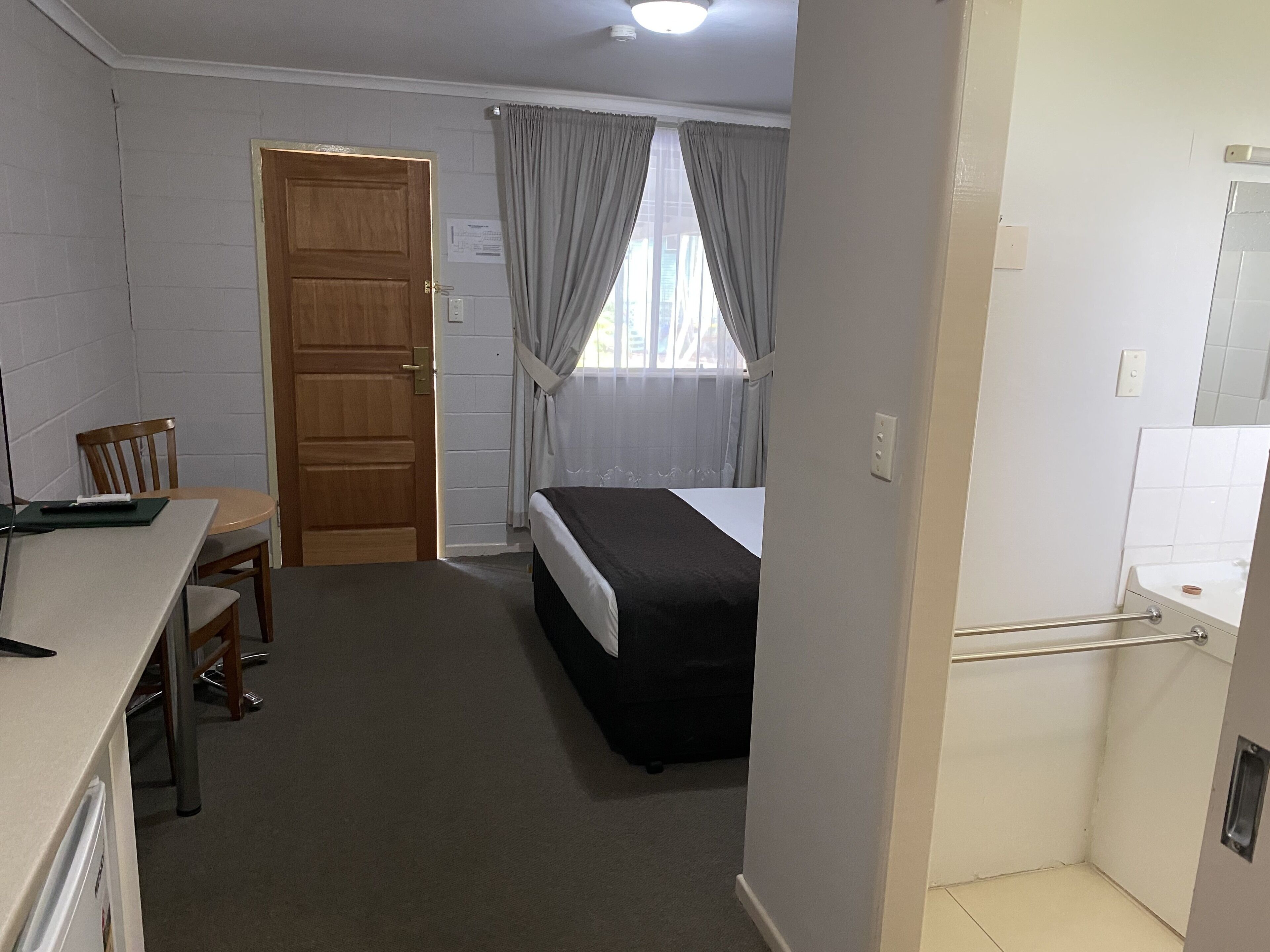 A small, clean room with a bed, a desk, and a chair. There is a window with curtains, a door, and a small bathroom area with a sink and mirror.