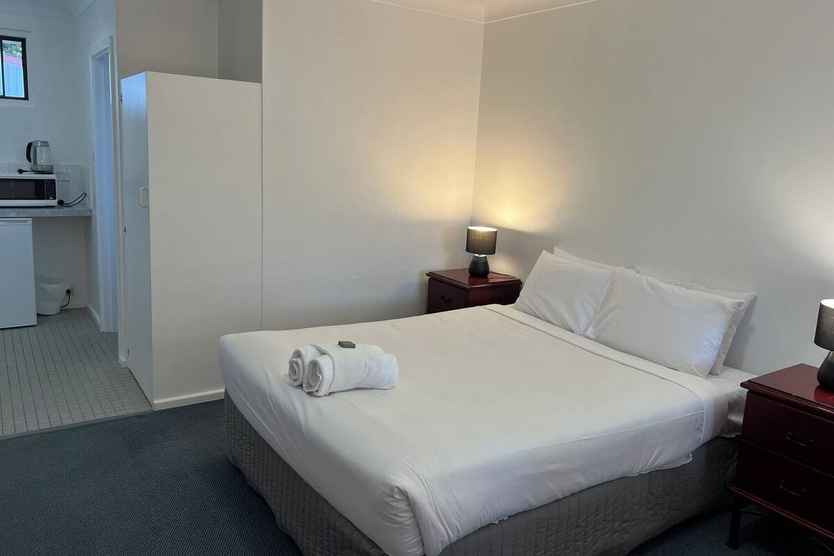 A hotel room with a neatly made bed, a bedside lamp, a small wooden nightstand, and a microwave in the corner.