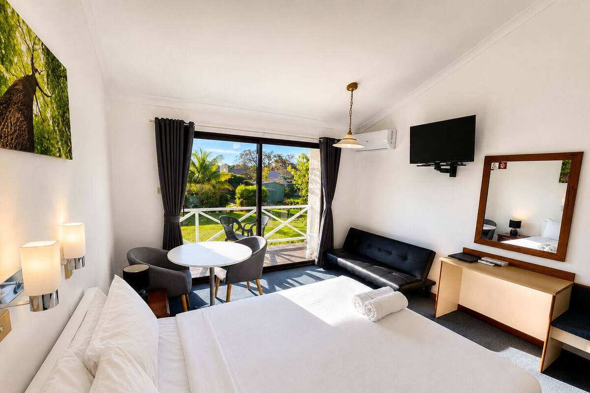 A hotel room with a bed, a desk, a TV, a balcony with a view of greenery, and a dining area.