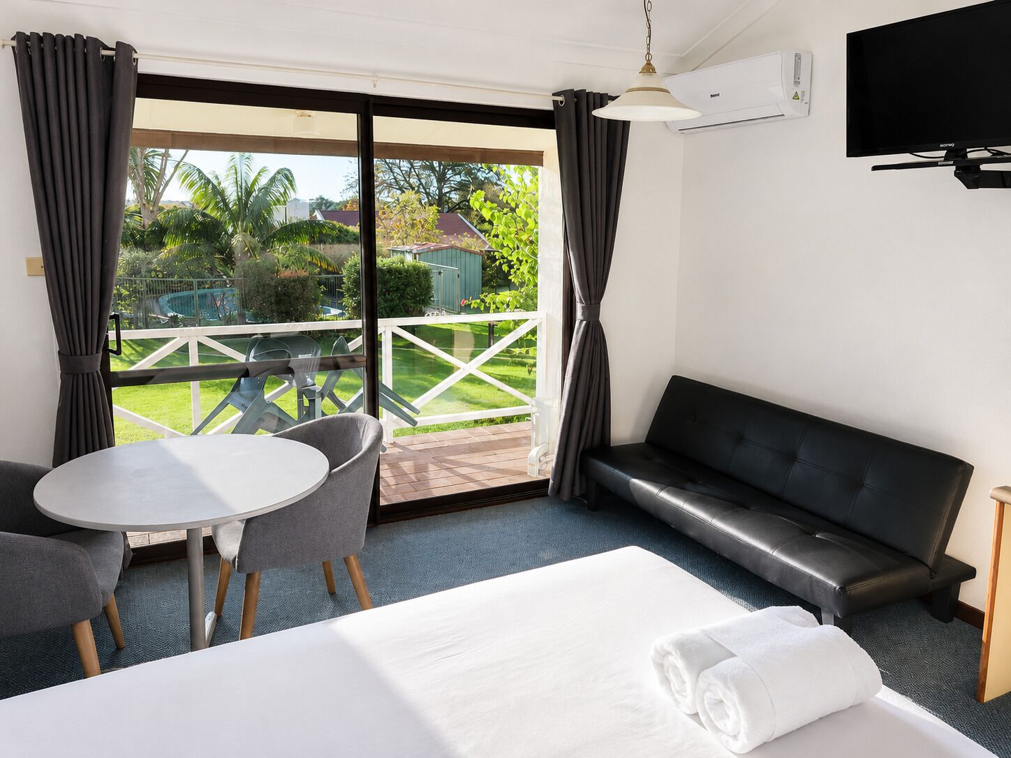 A hotel room with a bed, a round table with chairs, a black leather sofa, and a TV mounted on the wall.