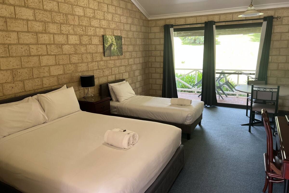 A hotel room with two beds, a small table, a chair, and a view of greenery through the window.
