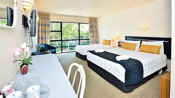 A hotel room with two beds, a dining table, chairs, a small table with a vase of flowers, and a window with curtains. - Manukau Hotel & Conference Centre (Auckland)
