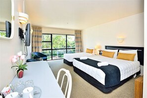 A hotel room with two beds, a dining table, chairs, a small table with a vase of flowers, and a window with curtains.