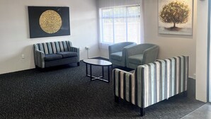 A modern lounge area with a dark carpet, a sofa, a striped armchair, and a small table. There are two windows with curtains, a ceiling with recessed lighting, and artwork on the walls. - Manukau Hotel & Conference Centre (Auckland)