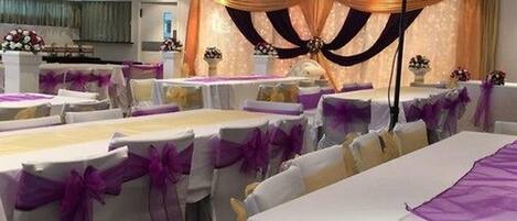 Un salón de banquetes con mesas redondas cubiertas con manteles blancos y cintas púrpuras, dispuestas en filas.