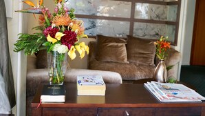 Una mesa de madera con un libro y periódicos, un jarrón con flores, y un sofá con cojines en una habitación con una pared de mármol y una ventana.