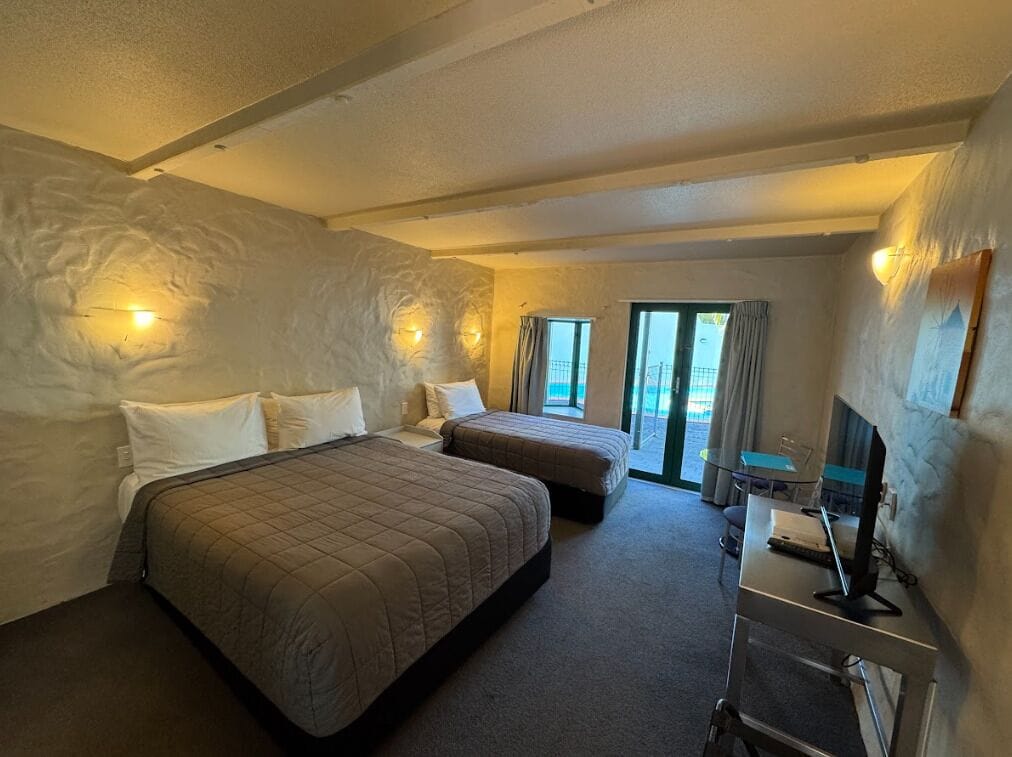 A hotel room with two beds, a desk with a computer, and a view of the outdoors.