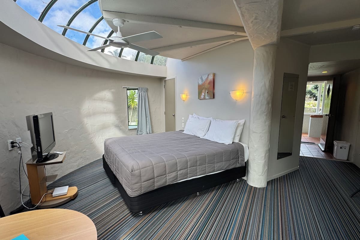 Un dormitorio moderno con techo curvo, una cama, un escritorio, un televisor y una ventana con cortinas.