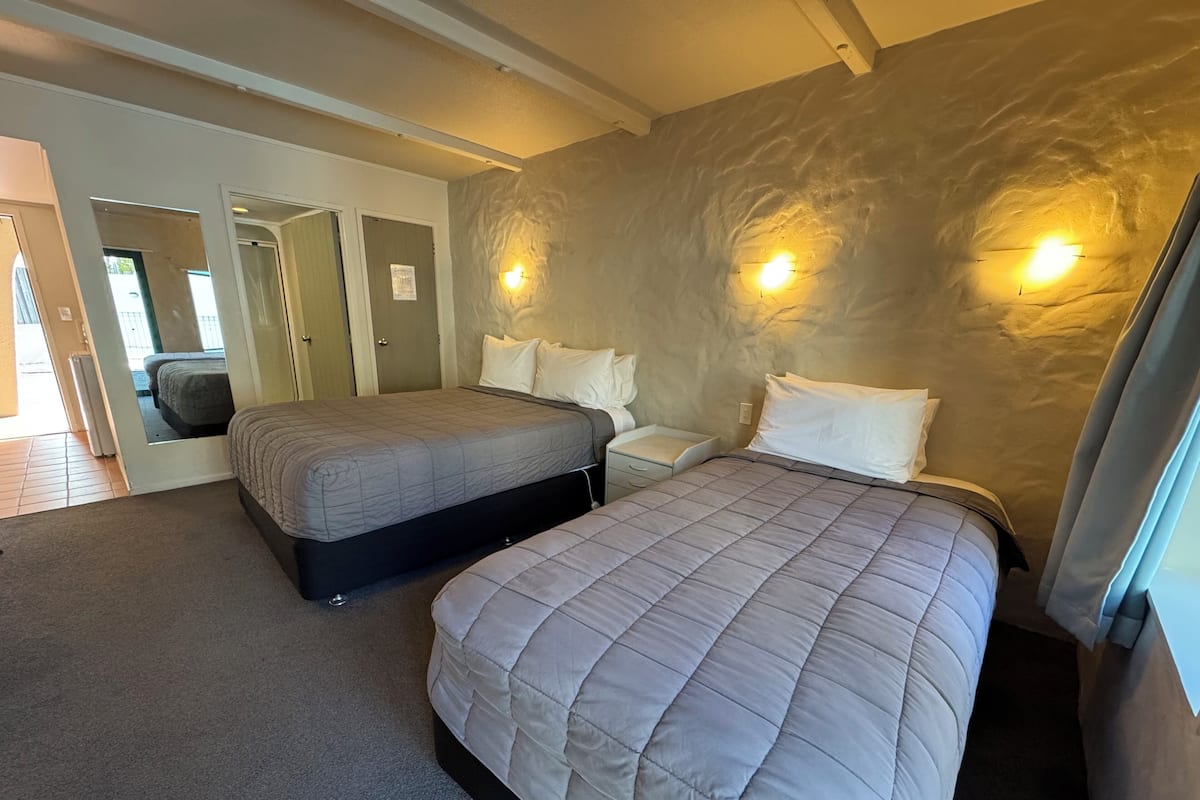 Un dormitorio con dos camas, una mesita de noche, una lámpara de pared y una puerta que da a otra habitación.