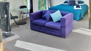 Una habitación moderna con techo curvo, una cama con colcha azul, un sofá púrpura, una mesita con una silla y un televisor.