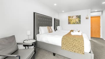 Una habitación de hotel moderna con una cama grande, un sofá gris, una mesita de noche con una lámpara y una puerta de madera.