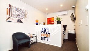 Ein Empfangsbereich mit einem Schild mit der Aufschrift 'AKL Airport Motel', einem schwarzen Sessel, einem kleinen Tisch mit einer Lampe und einem an der Wand befestigten Fernseher.