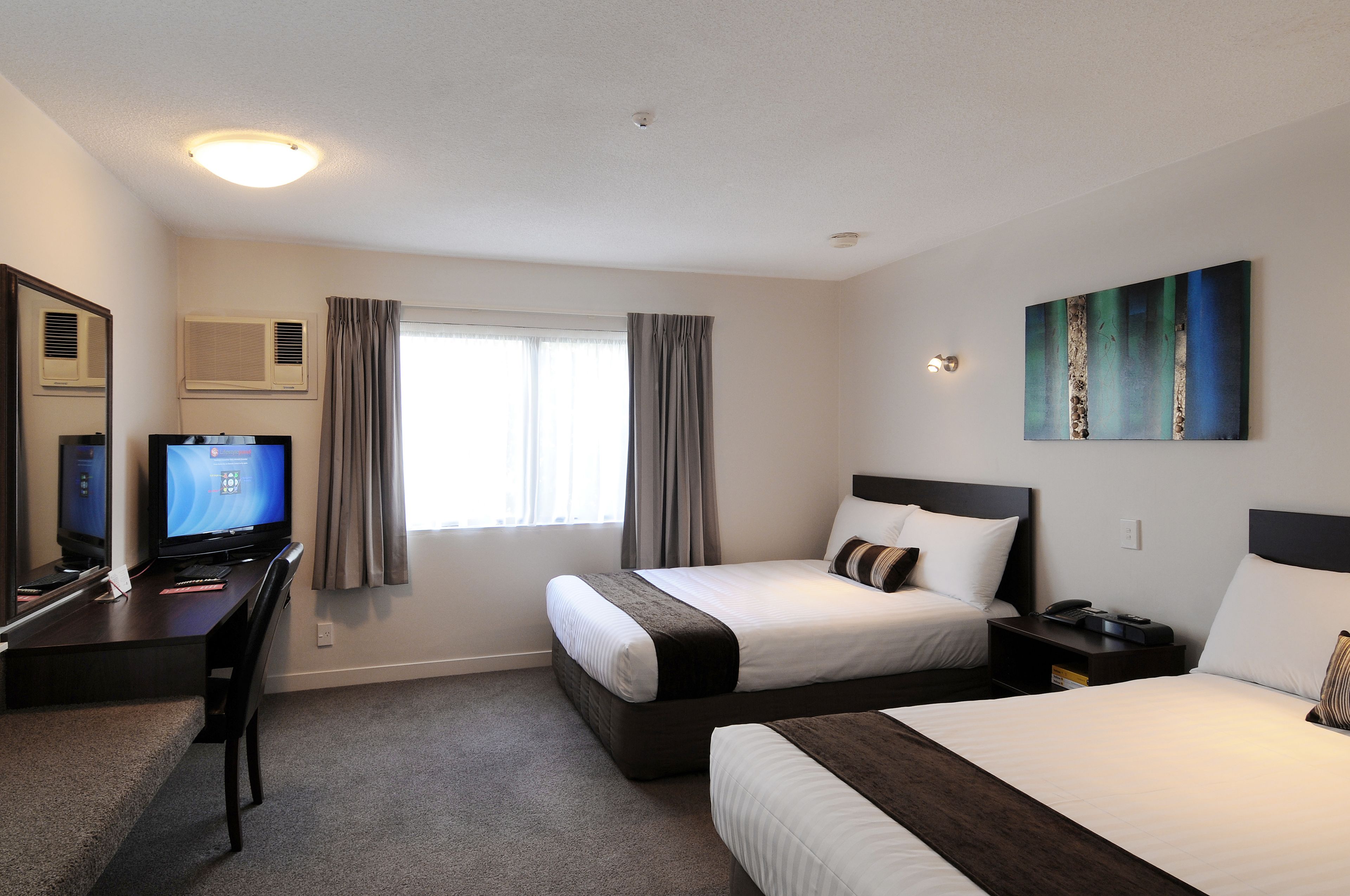 A hotel room with two beds, a desk with a computer, a window with curtains, and a painting on the wall.