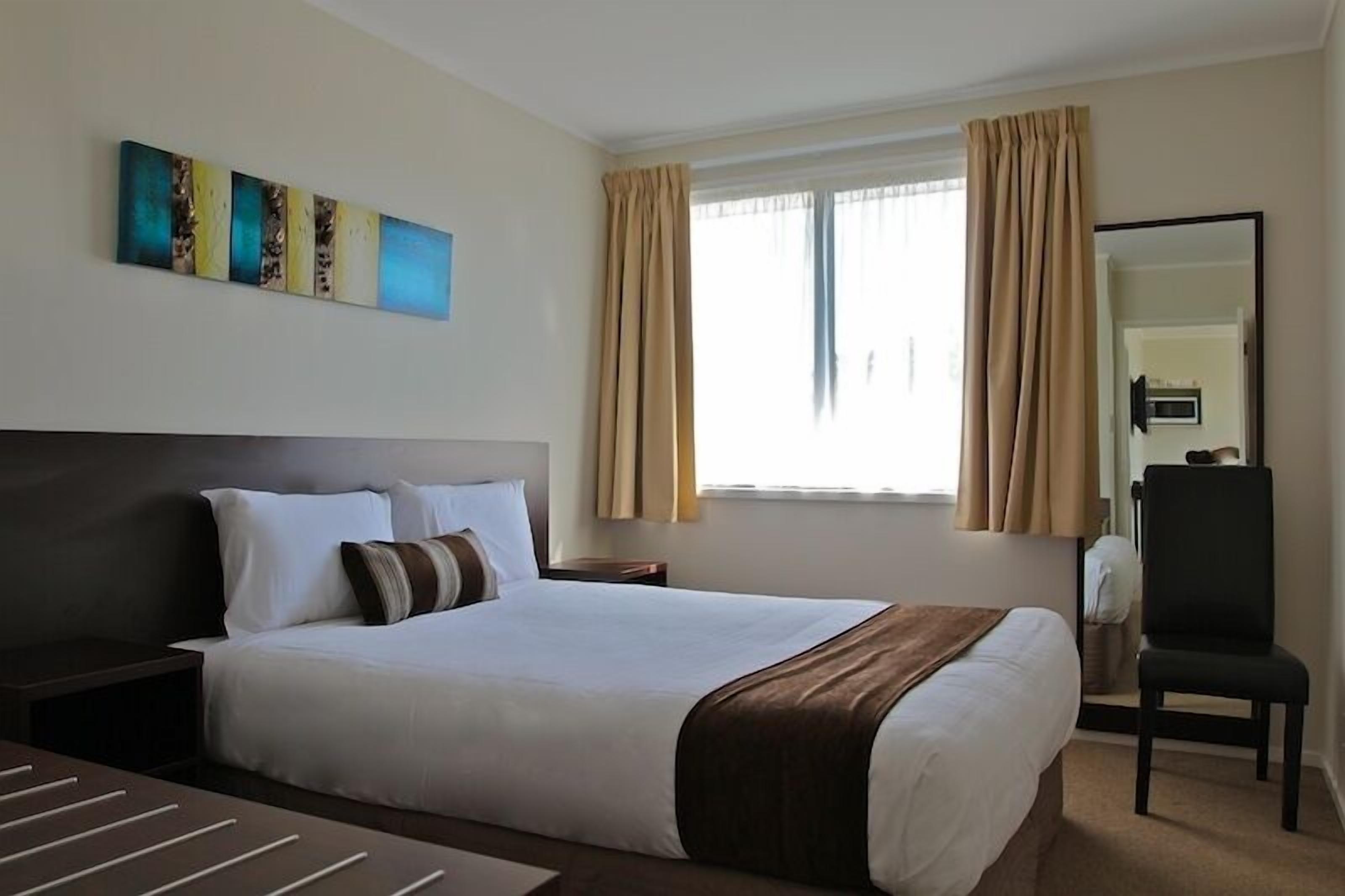 Une chambre d’hôtel avec un grand lit, une table de chevet, une chaise, une fenêtre avec des rideaux et un tableau au mur.