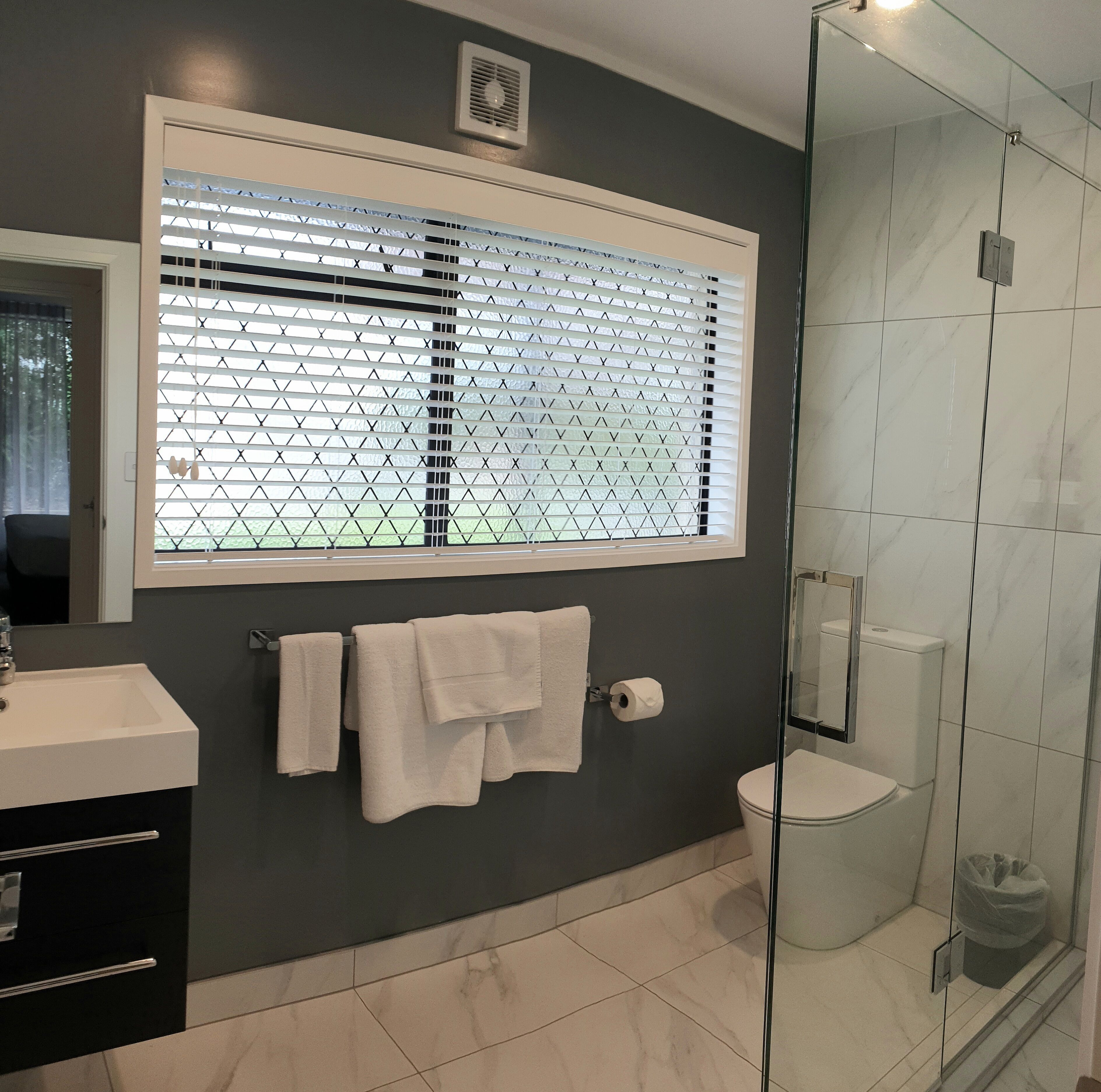 Un baño con cabina de ducha de vidrio, inodoro, lavamanos con espejo y ventana con persianas.